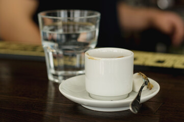 a finished white cup of coffee with a white saucer and spoon on the blurred background of a glass glass with water
