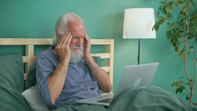 A Retired Elderly Man Working At Home Feels Stress, Headache While Working With A Computer While Lying In Bed At Home. Modern Technology And Health Concept.