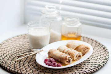 pancakes rolled up on a white plate with frozen raspberries and honey on the background of a glass of milk, a jar of honey and sugar on a wicker napkin