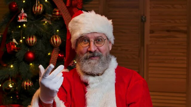 Portrait of Santa Claus looking at the camera waves hello and makes gesture come here. Old man in red suit and hat against the background of Christmas tree with toys and lights. Close up. Slow motion.