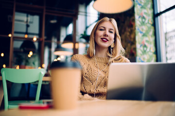 Smiley woman using laptop and earphones at cafe table with coffee and smartphone