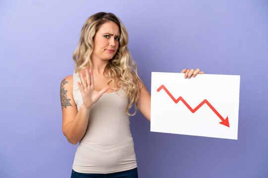 Young Brazilian woman isolated on purple background holding a sign with a decreasing statistics arrow symbol and doing stop sign