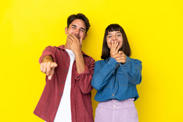 Young mixed race couple isolated on yellow background pointing with finger at someone and laughing a lot
