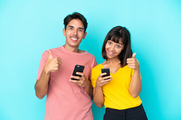 Young mixed race couple holding mobile phone isolated on blue background giving a thumbs up gesture with both hands and smiling