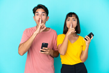 Young mixed race couple holding mobile phone isolated on blue background showing a sign of silence gesture putting finger in mouth