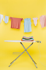 stack of clean clothes on ironing board under laundry hanging on rope on yellow background.