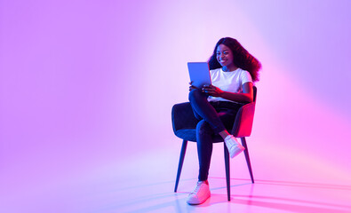 Cheerful black lady using tablet, studying or working remotely, sitting in armchair in neon light,...