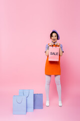 astonished woman showing shopping bag with sale lettering near purchases on pink.