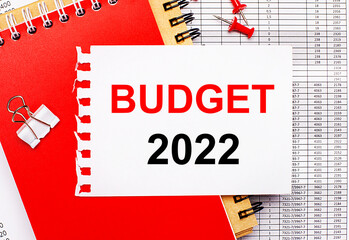 Against the background of the reports, there are red and brown notepads, a white paper clip, red buttons and a white sheet of paper with the text BUDGET 2022. Business concept