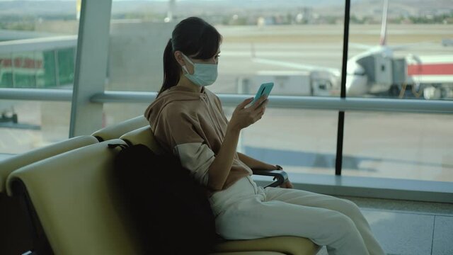 Woman in medical mask waiting boarding in big empty airport. Female traveler look at phone, search for boarding pass or flight cancelation information.