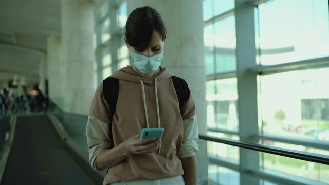 Young woman in protective face mask in big empty airport. Female traveler look at phone, search for boarding pass or flight cancelation information.