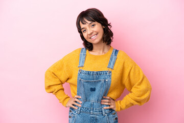 Young woman with overalls isolated background posing with arms at hip and smiling - obrazy, fototapety, plakaty