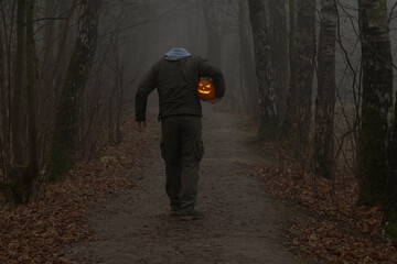 Headless man holds Jack O'Lantern Halloween pumpkin head under his arm and walks on countryside road in autumn foggy forest. Carved face on the pumpkin glows orange. Halloween costume theme.