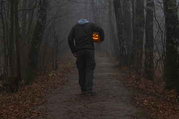 Headless man holds Jack O'Lantern Halloween pumpkin head under his arm and walks on countryside road in autumn foggy forest. Carved face on the pumpkin glows orange. Halloween costume theme. - obrazy, fototapety, plakaty