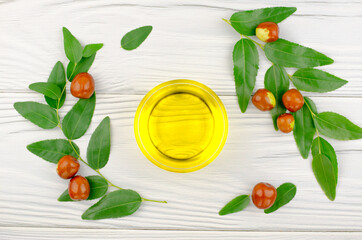 Jojoba oil in a bowl, a sprig of jojoba and fruits on a white wooden table - obrazy, fototapety, plakaty