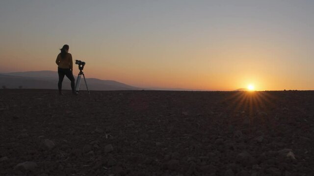 Low angle wide view of a woman filming the sunset with camera and tripod in the desert. Slow motion. 
