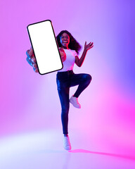 Full length of excited black woman jumping with cellphone, showing blank screen in neon light,...