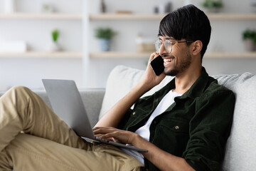 Cheerful indian guy independent contractor with laptop having phone conversation