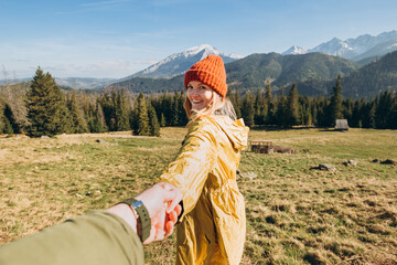 Hiker with red hat stands on edge of cliff against background and extends a hand. Happy woman gives a hand to someone like follow me. First person view.