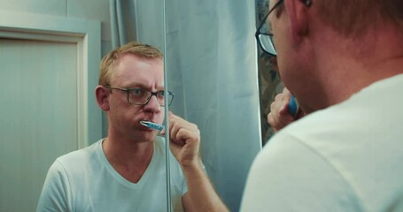Head shot mirror reflection man in glasses brushing teeth with toothbrush and toothpaste. Morning oral anti cavity hygienic routine alone in bathroom