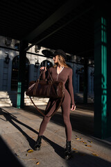 Fototapeta na wymiar Fashionable young brunette woman with long legs wearing Solid Long Sleeve Bodycon One Piece Jumpsuits posing with a leather brown travel bag