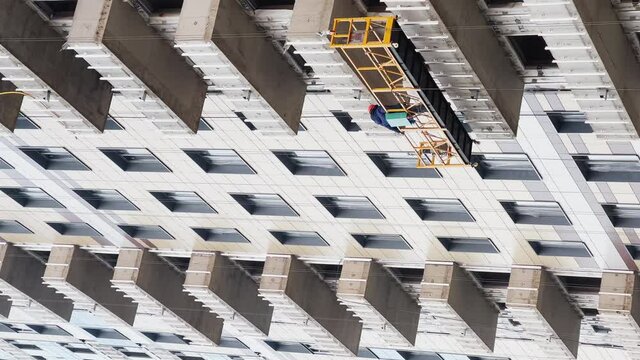 Construction of the high floor house, The worker in a protective helmet does a facade, construction cranes, the bottom view up, concrete blocks with windows, cloudy weather, a huge real estate