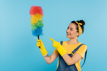 smiling housewife in yellow rubber gloves pointing at colorful dust brush isolated on blue.