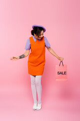 full length view of amazed woman looking at shopping bag with sale lettering on pink.