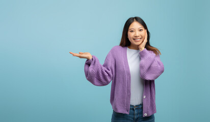 Wow, cool offer. Cheerful asian lady showing empty space on palm and touching cheek standing over blue background