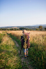 Little hiker with backpack comes from the mountain and carries a bouquet of wildflowers that he collected for his mother, path among wheat, gorgeous landscape, beauty of nature, view from behind