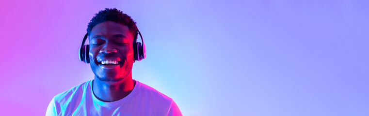 Handsome young black man wearing headphones, listening to music with closed eyes in neon light,...