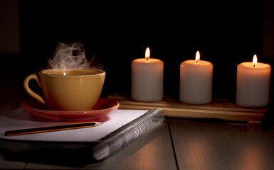Fototapeta na wymiar Cup of hot coffee with yellow smoke, notebook with pencil and burning candles. Desk table in power supply cut