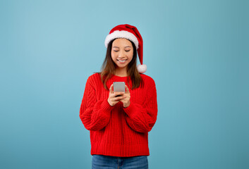 Excited asian lady in santa hat looking at smartphone, texting xmas greeting message, standing over blue background