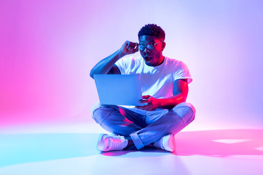 Shocked black guy sitting cross legged with laptop, touching his glasses, winning online lottery in neon light