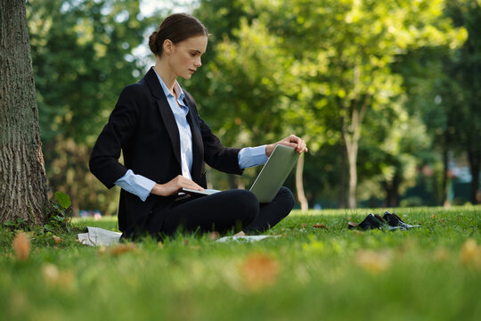 Side view of woman sitting in a park on green grass with a lapot and notebook, outdoor worker, remote work.