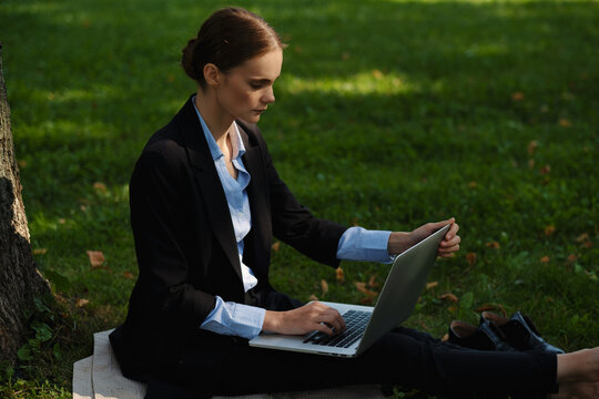 Side view of woman sitting in a park on green grass with a lapot and notebook, outdoor worker, remote work.