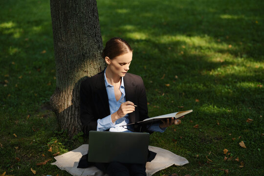 Side view of woman sitting in a park on green grass with a lapot and notebook, outdoor worker, remote work.