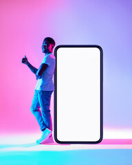 Happy black guy leaning on big cellphone with empty screen, showing thumb up, recommending new app in neon light, mockup