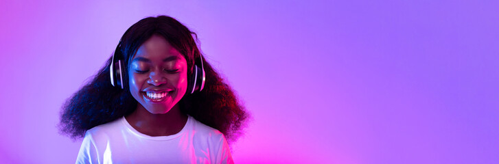 Pretty young black woman in headphones listening to music with closed eyes in neon light, panorama