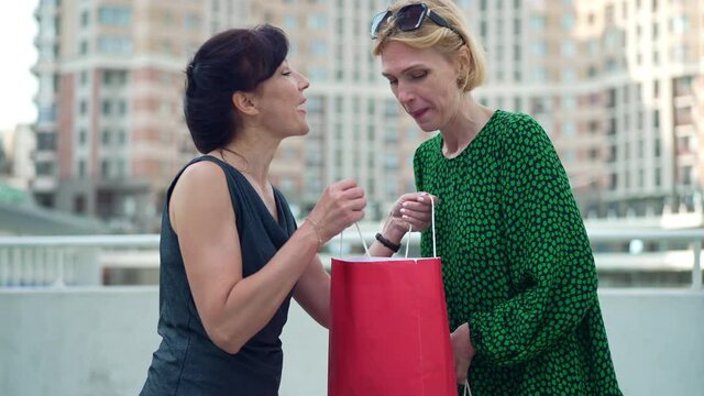 Two envious adult women bragging purchases in shopping bags talking standing on city street. Portrait of Caucasian jealous friends showing off boasting to each other outdoors. Slow motion