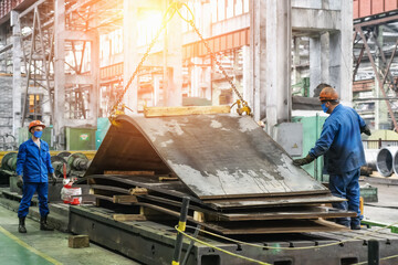 Fototapeta premium Workers in masks and helmets at metallurgical plant unload steel or metal sheets with crane for steelmaking production.