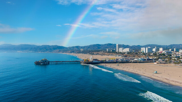 a stunning aerial shot of the coastline with silky brown sand, vast blue ocean water, a long wooden pier with carnival rides, beachfront buildings, clouds in a blue sky, rainbow at Santa Monica Beach	