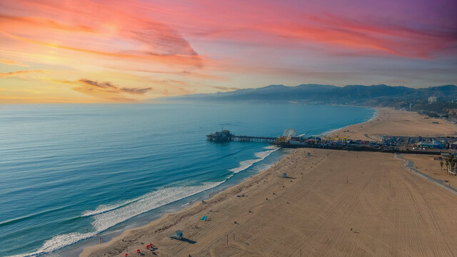 a stunning aerial shot of the coastline with silky brown sand, vast blue ocean water, a long wooden pier with carnival rides, buildings, clouds at sunset at Santa Monica Beach in California USA