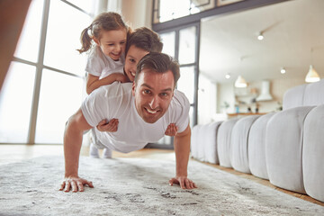 Portrait of smiling father exercising while holding daughter and son on his back during workout indoors - obrazy, fototapety, plakaty