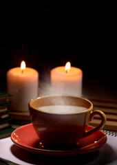 Hot yellow mug with drink, steam and two burning candles together on the table, black background space for text.