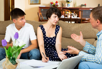 Focused woman with teenage son discussing apartment rental agreement with landlord, signing papers on sofa at home