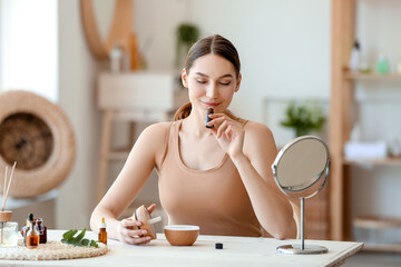 Young woman sitting at table and smelling natural essential oil in bottle - obrazy, fototapety, plakaty