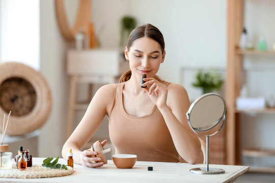 Young Woman Sitting At Table And Smelling Natural Essential Oil In Bottle