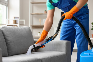 Male worker removing dirty stain from grey sofa with vacuum cleaner in room - obrazy, fototapety, plakaty