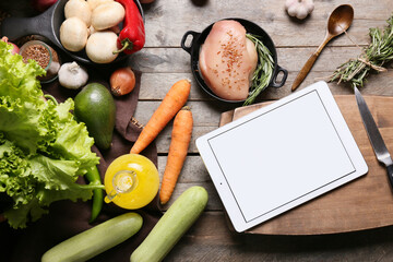 Tablet computer with fresh vegetables and raw chicken meat on dark wooden table
